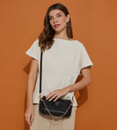 A woman models a black Prada shoulder bag with a gold chain strap and logo, paired with a cream top and beige trousers, set against a solid orange background.