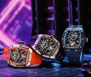 Three Richard Mille chronograph watches with skeletonized dials are displayed on a dark metallic surface. The watches feature orange, rose gold, and dark blue/gray cases with matching rubber straps, set against a futuristic purple and black background.