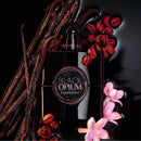 A black perfume bottle labeled 'Black Opium' by Yves Saint Laurent, styled with vanilla beans, coffee beans, and pink orchids on a glossy black background.