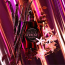 A perfume bottle labeled 'Black Opium' surrounded by vanilla pods, coffee beans, and pink orchid flowers against a dramatic purple and magenta striped background.