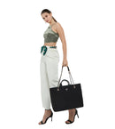 A woman modeling a black quilted Prada tote bag with gold-tone chain handles, styled with a green camouflage crop top and white wide-leg pants against a light gray studio background.