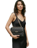 A woman wearing a black satin slip dress holds a black woven Guess shoulder bag with a gold-tone chain strap against a light gray studio background.