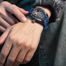 A close-up of a person's wrist wearing a skeleton-style watch with a dark blue strap and a black rectangular case, set against a denim jacket background.