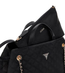 Close-up of a black GUESS handbag with quilted texture, gold-tone hardware, and a zippered pouch partially inserted into the main compartment. The GUESS logo is visible on the pouch and triangular metal badge on the bag.